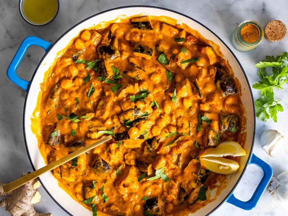 Eggplant curry in a blue skillet with a golden spoon and lemon.