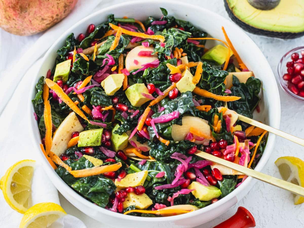 Kale salad in a white bowl with lemons and avocado on the side