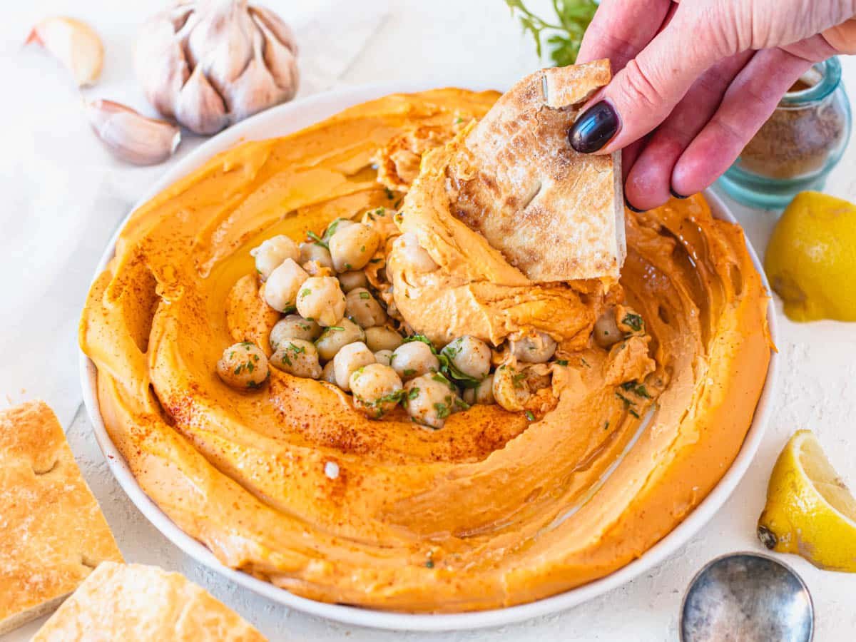 roasted pepper hummus in a white bowl with hand holding a pita bread