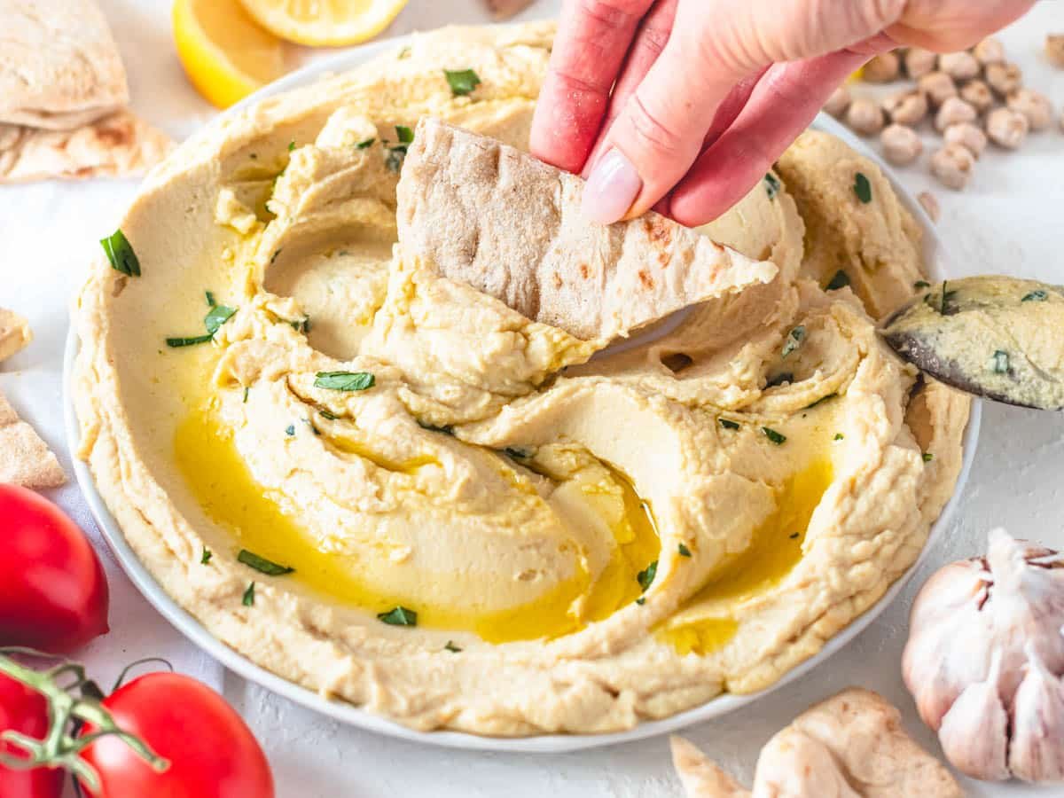 Hummus without tahini on a plate with hand holding a pita chip
