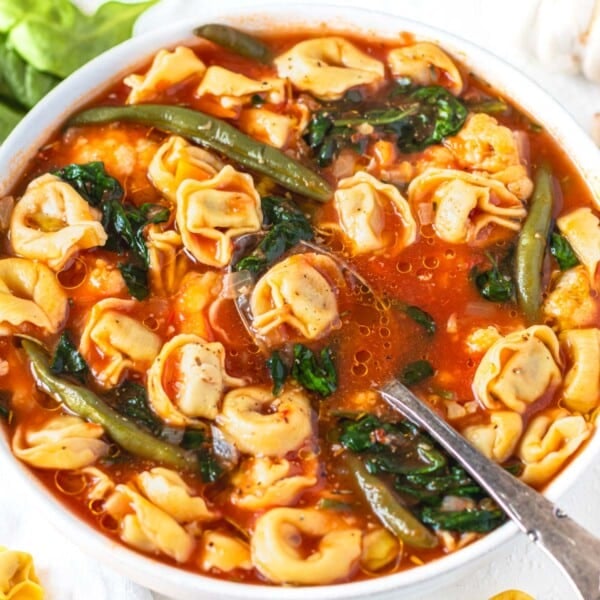 Tortellini soup in a white bowl with silver spoon and greens