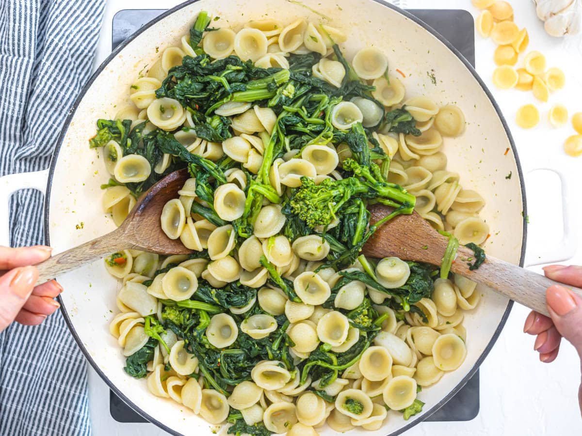 orecchiette and broccoli rabe mixing in a white skillet with hands