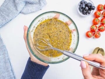 Greek dressing in a glass bowl with hand holding a whisk