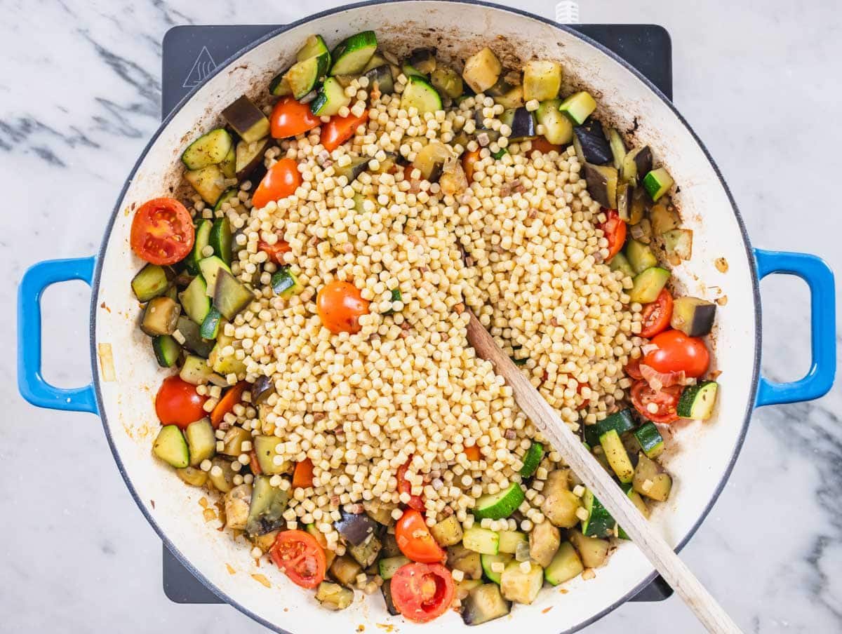 Fregola added to vegetables in a skillet with a wooden spoon