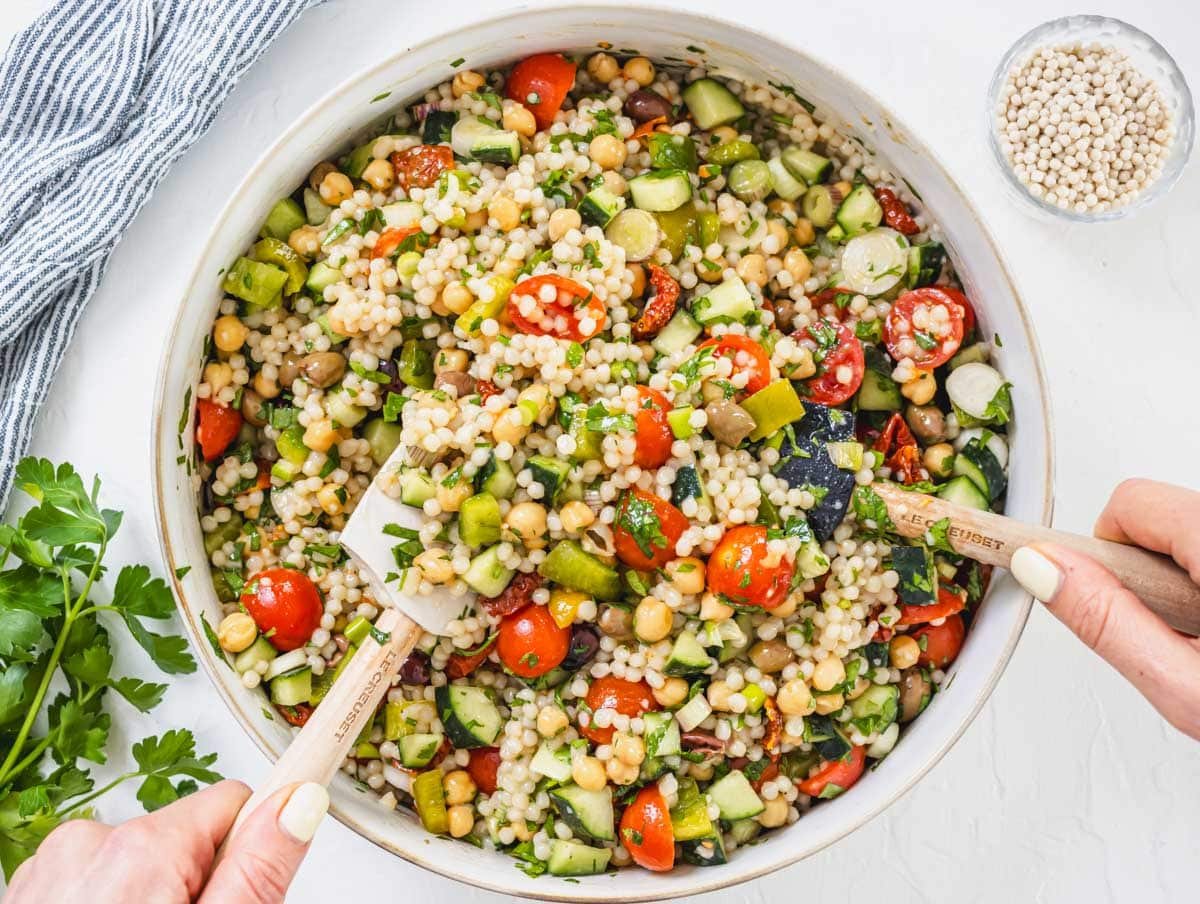 hands mixing pearl couscous and fresh ingredients in a bowl
