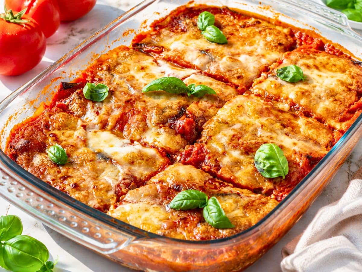 Freshly baked Eggplant Parmigiana cooling beside serving basil leaves on white marble countertop.