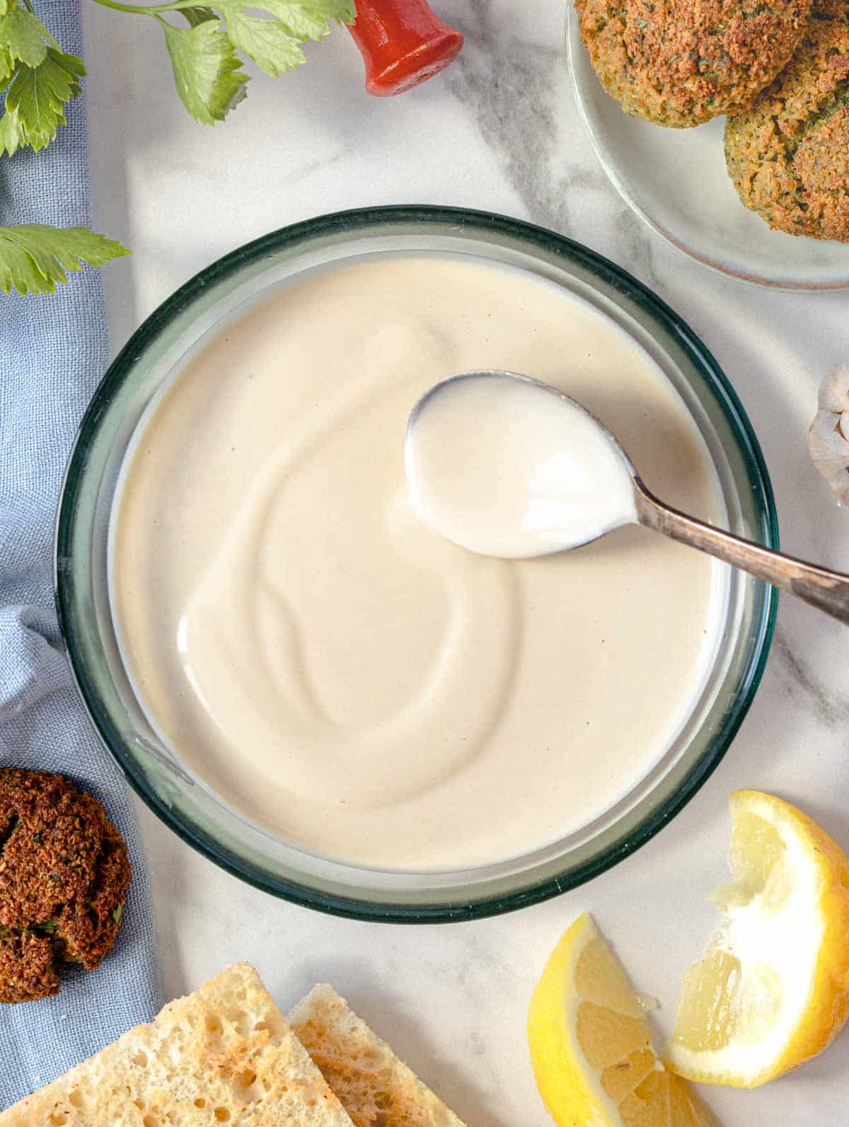 Tahini sauce in a glass bowl with a small silver spoon and lemon wedges
