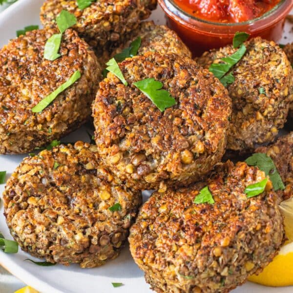 Golden lentil patties with visible lentils stacked with fresh parsley and lemon on a white plate.