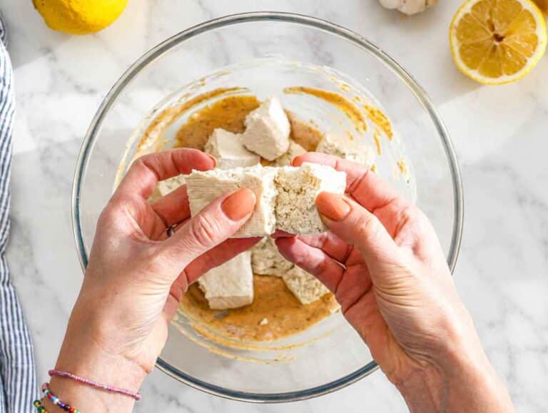 Step 2 Hands tearing firm tofu into rustic bite-sized pieces over white marble.