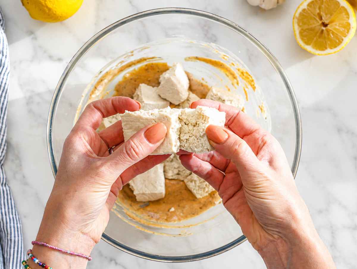 Step 2 Hands tearing firm tofu into rustic bite-sized pieces over white marble.
