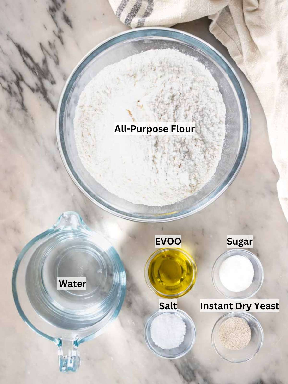 Ingredients for focaccia dough including flour, yeast, olive oil, salt, and water on table.