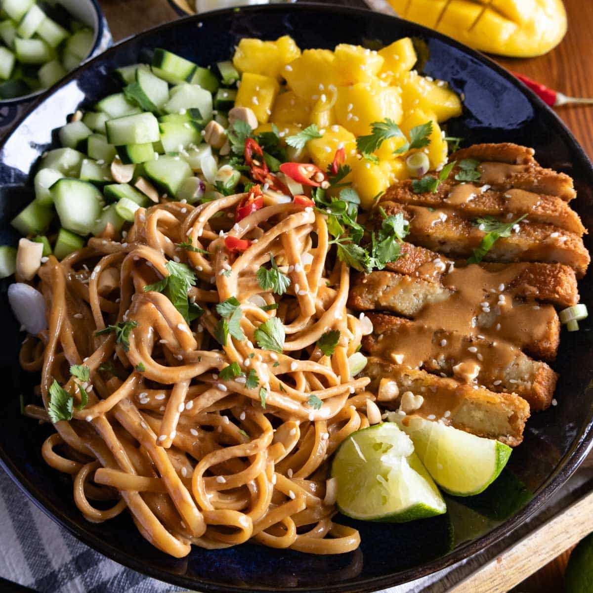 Sesame noodles in a dark bowl topped with mango, cucumber, and lime.