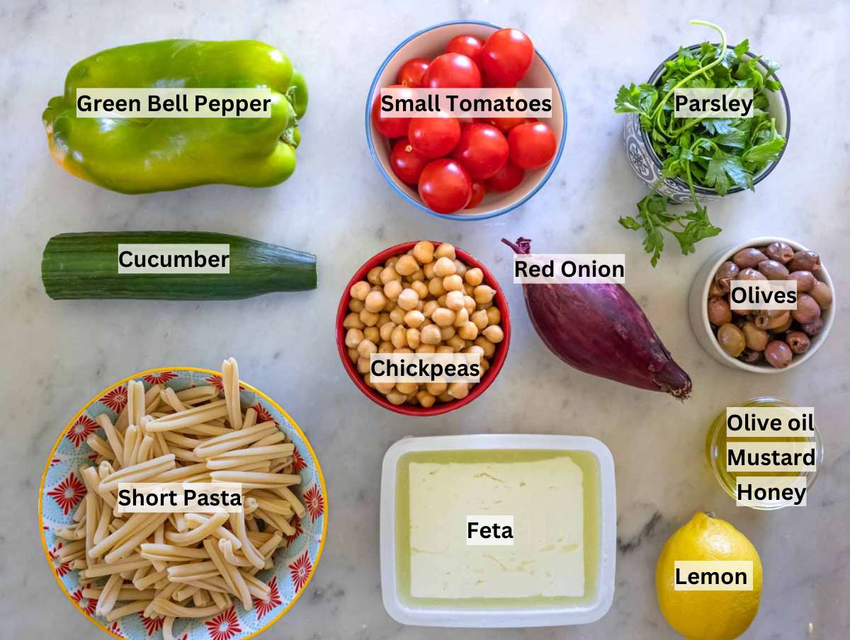 Ingredients for Greek pasta salad including chickpeas, pasta, cherry tomatoes, cucumber, red onion, feta, olives, herbs, and olive oil dressing.