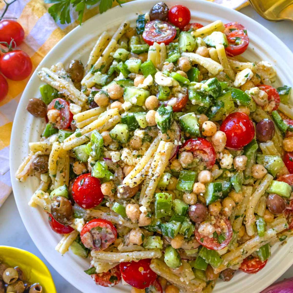 Platter of Greek pasta salad with feta, olives, cucumber, and tomatoes served with lemon wedges – easy Mediterranean summer recipe.