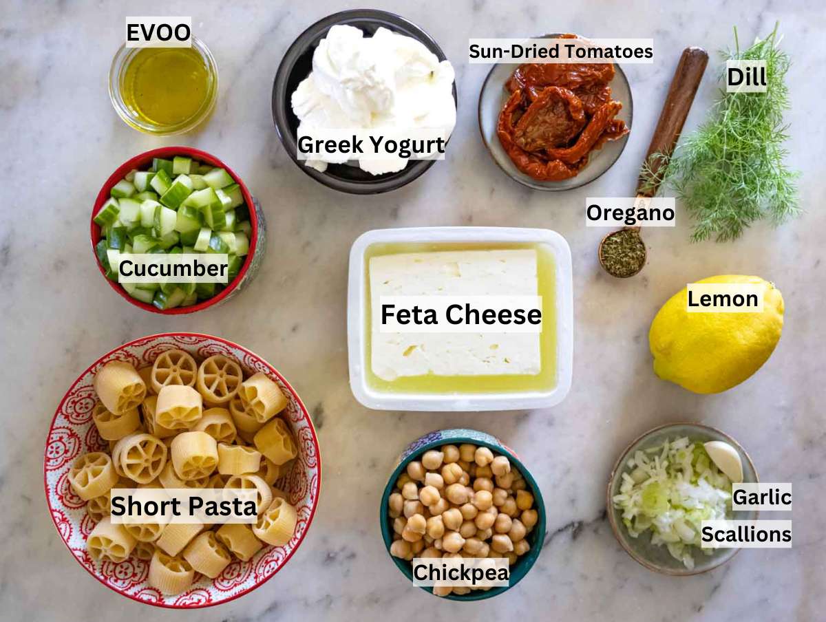 All ingredients for cold pasta salad laid out - pasta, yogurt, feta, chickpeas, cucumber, herbs
