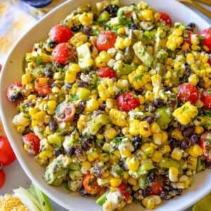 Colorful corn salad with black beans, avocado, and lime dressing served in a white bowl, styled with fresh ingredients around it—ideal vegetarian BBQ side dish.