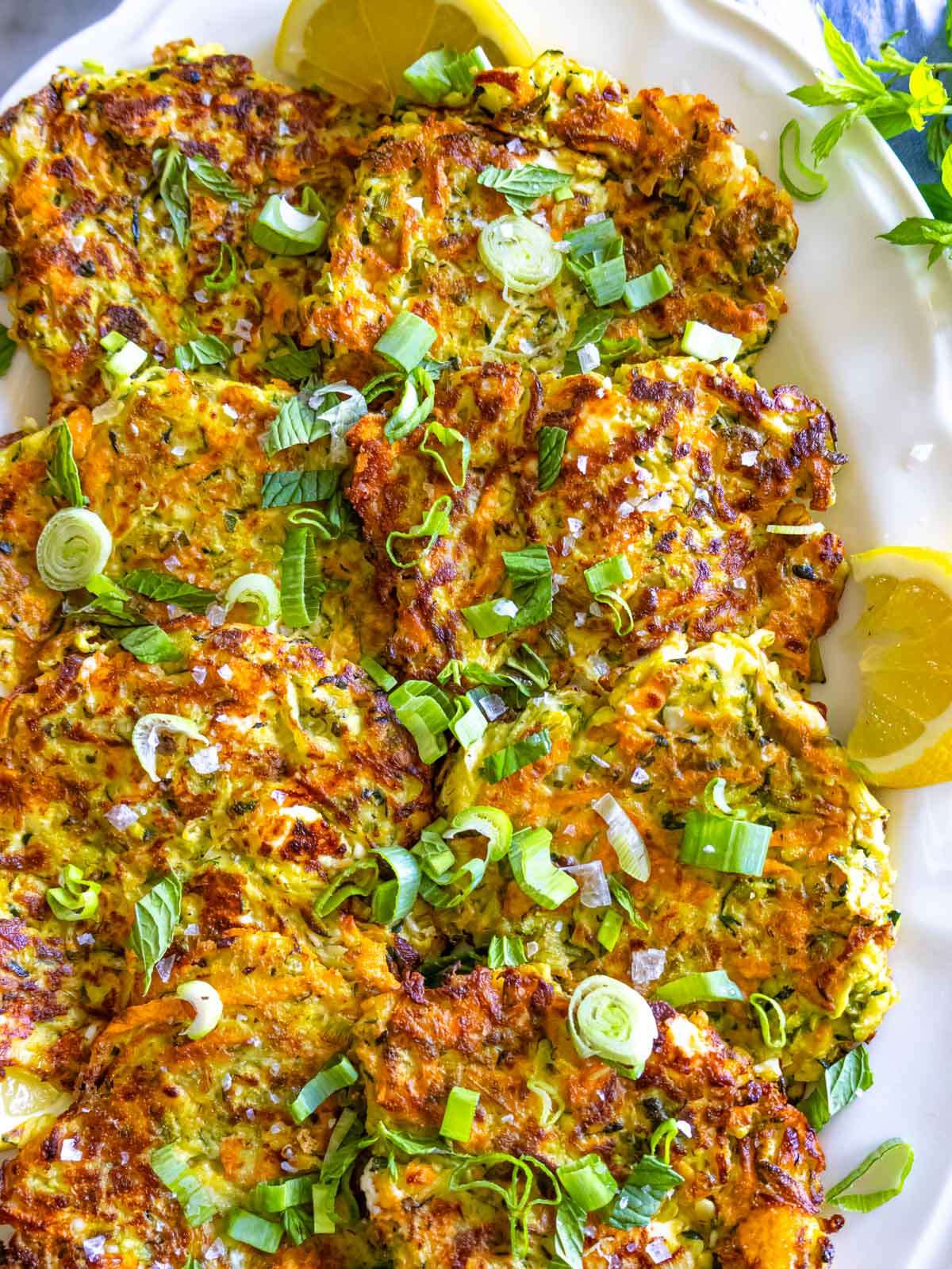 Crispy zucchini patties with golden edges and fresh herbs, served with yogurt sauce on a white plate.