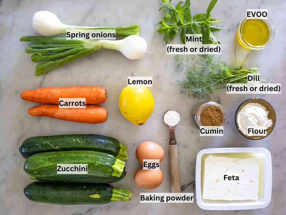 Flat lay of fresh ingredients for zucchini patties including zucchini, carrots, eggs, mint, dill, feta, lemon, and flour on a wooden table.
