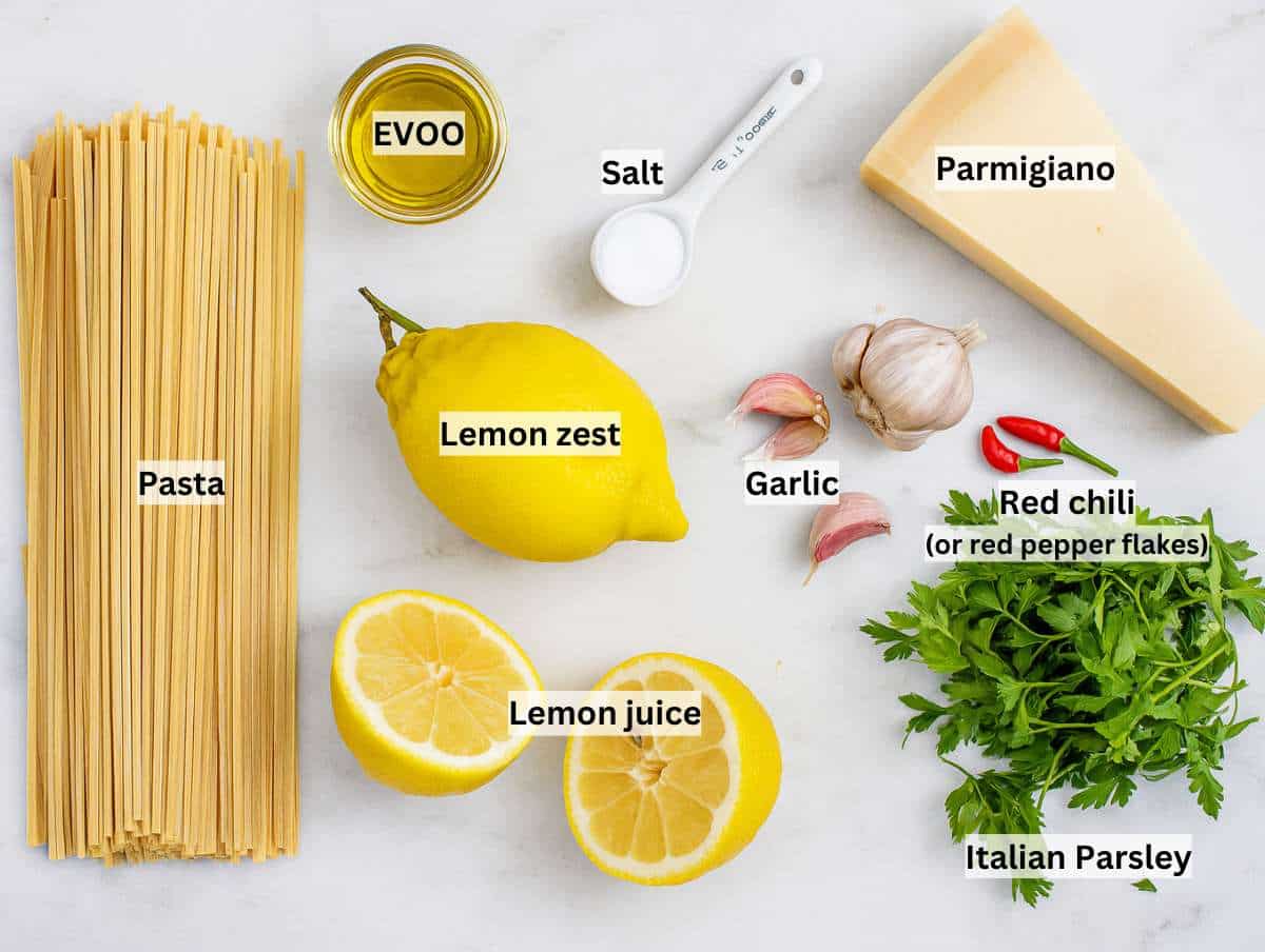 Flat lay of ingredients for lemon pasta without cream, including spaghetti, garlic, lemon, olive oil, parsley, chili, and parmesan cheese.