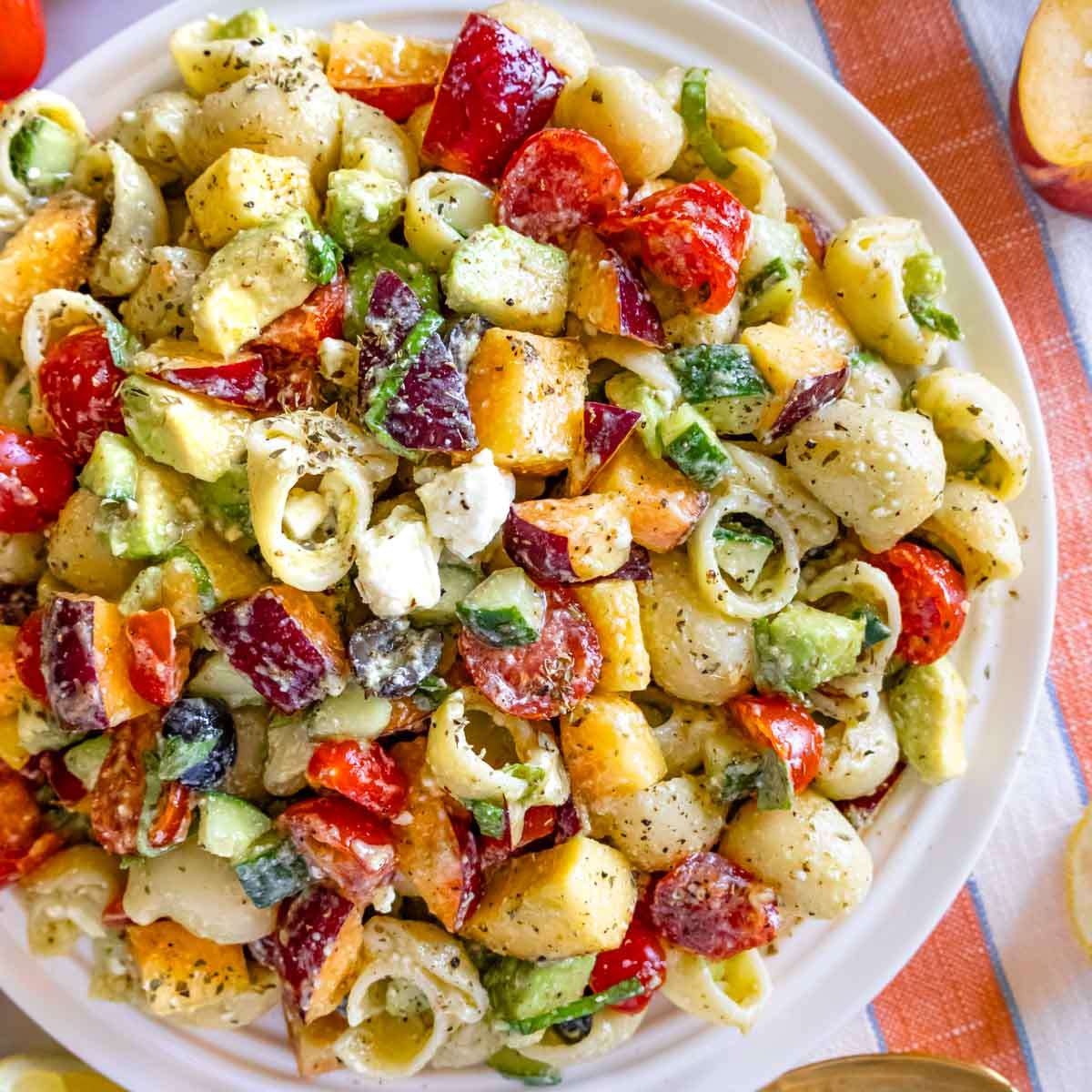 Fresh and colorful Mediterranean peach pasta salad with avocado, tomatoes, and olives on a summer table with basil leaves and lemon in the background.