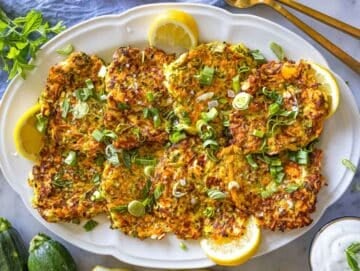 Freshly cooked zucchini patties served on a platter with fresh herbs and lemon wedges.