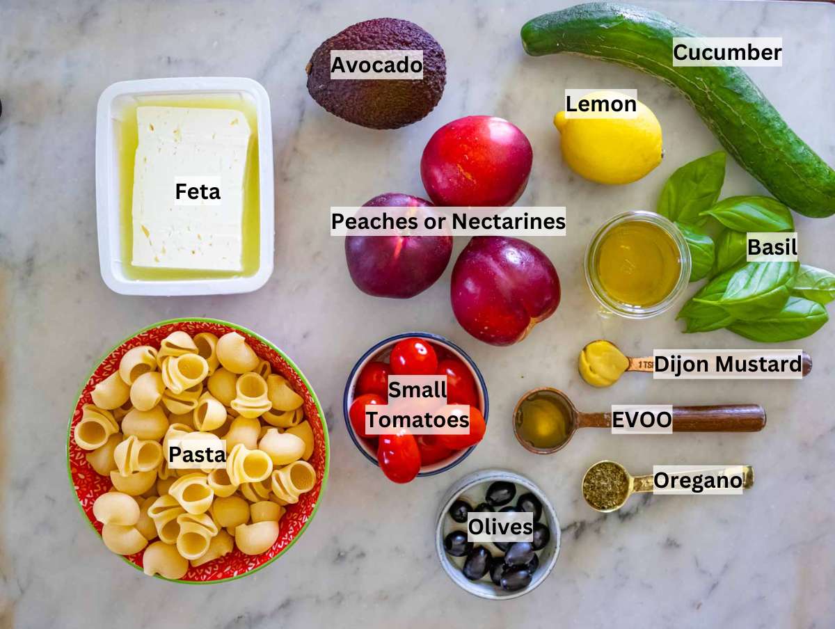 Ingredients laid out for summer pasta salad, including peaches, feta, avocado, pasta, cucumber, cherry tomatoes, olives, and lemon.