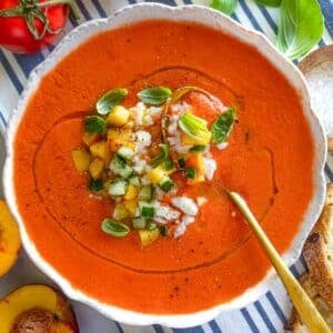 Peach gazpacho in a bowl with spoon, fresh peaches and basil in the background.