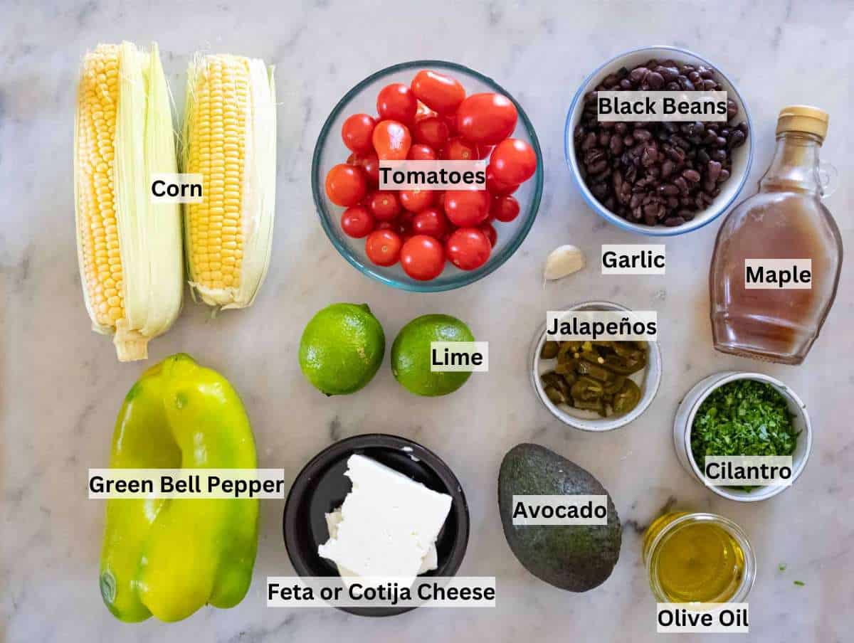 Raw ingredients for corn salad including fresh corn on the cob, black beans, avocado, cherry tomatoes, red onion, green bell pepper, cilantro, lime, and feta arranged on a marble surface.