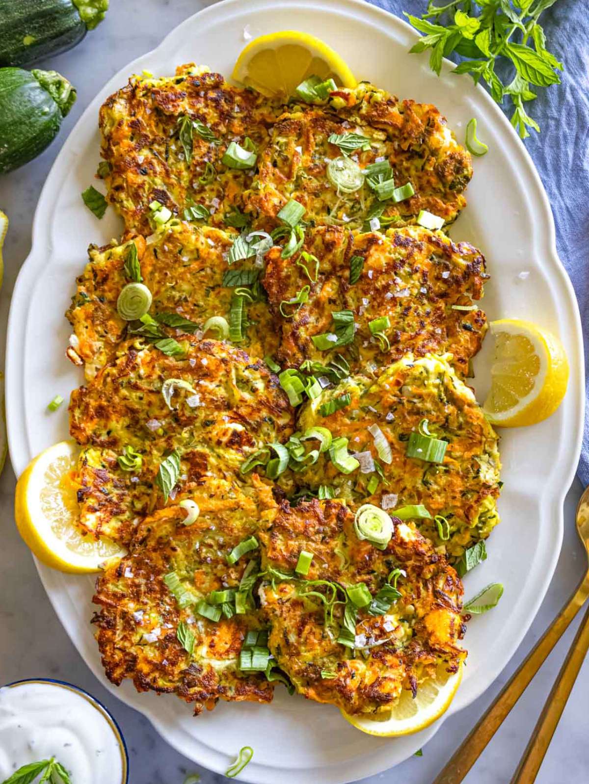 Top-down view of Greek-style zucchini patties arranged on a platter with lemon wedges, dill, and a side of tzatziki.