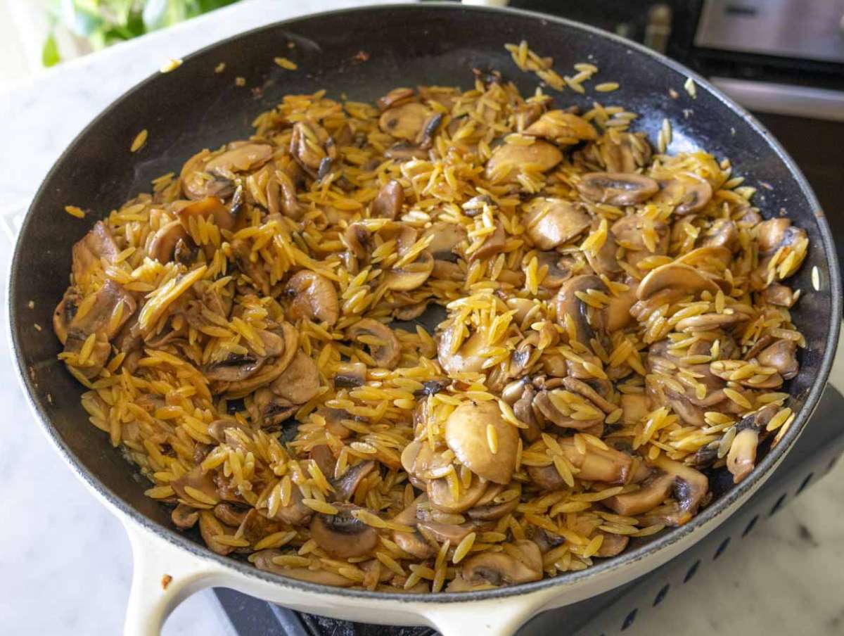 Dry orzo pasta being stirred into mushrooms in pan before adding broth.