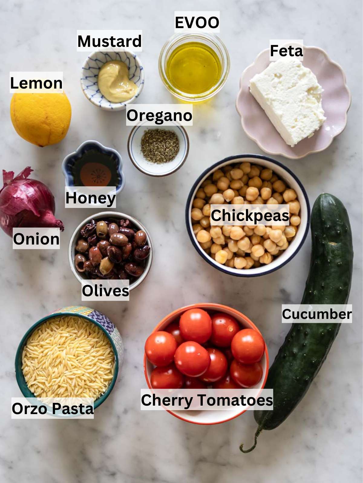 Fresh ingredients for Greek Orzo Salad including orzo pasta chickpeas tomatoes cucumber feta and olives.