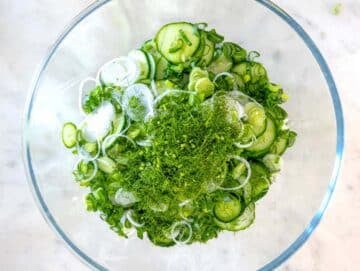 Sliced cucumbers green onion and fresh dill in a mixing bowl ready for cucumber salad.