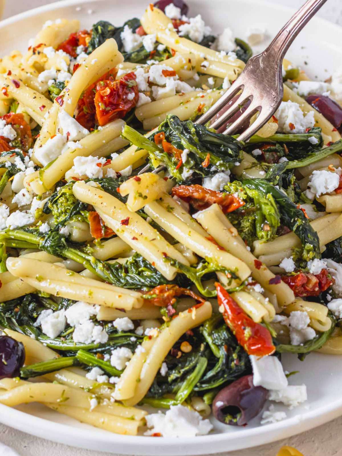 Close up of Mediterranean pasta with broccoli rabe olives and feta in a white bowl.