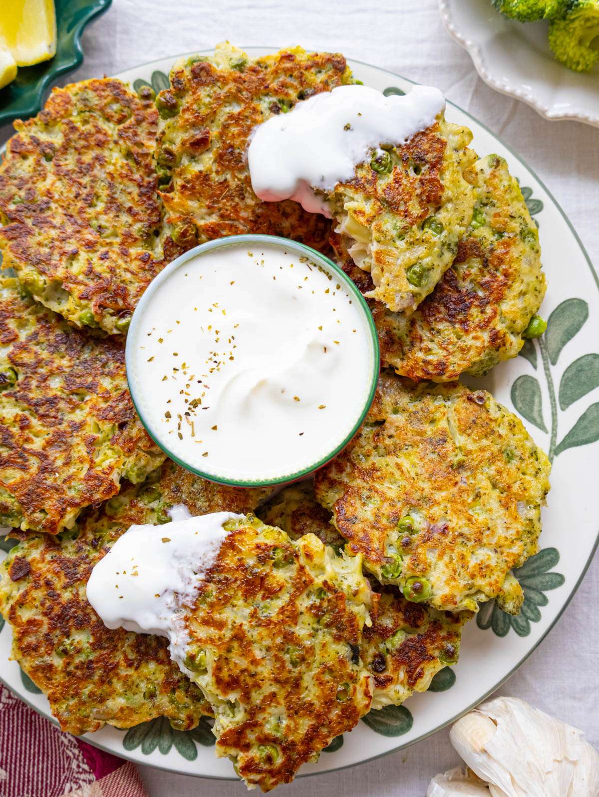 Top-down view of broccoli fritters on a plate with yogurt dip and lemon wedges on the side.