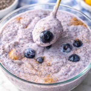 Bowl of blueberry chia pudding on a table with spoon and scattered blueberries in natural light.