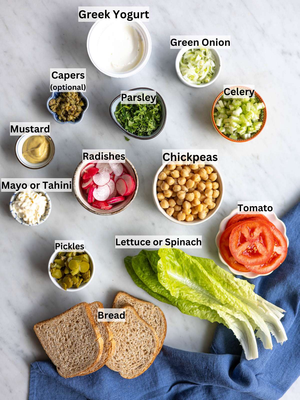 Bowls of chickpeas, yogurt, chopped vegetables, herbs, mustard, and lemon juice arranged on a marble table.