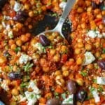 Chickpea shakshuka ready in a skillet topped with crumbled feta, oregano, parsley, and olives.