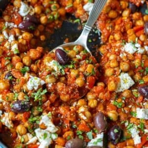 Chickpea shakshuka ready in a skillet topped with crumbled feta, oregano, parsley, and olives.