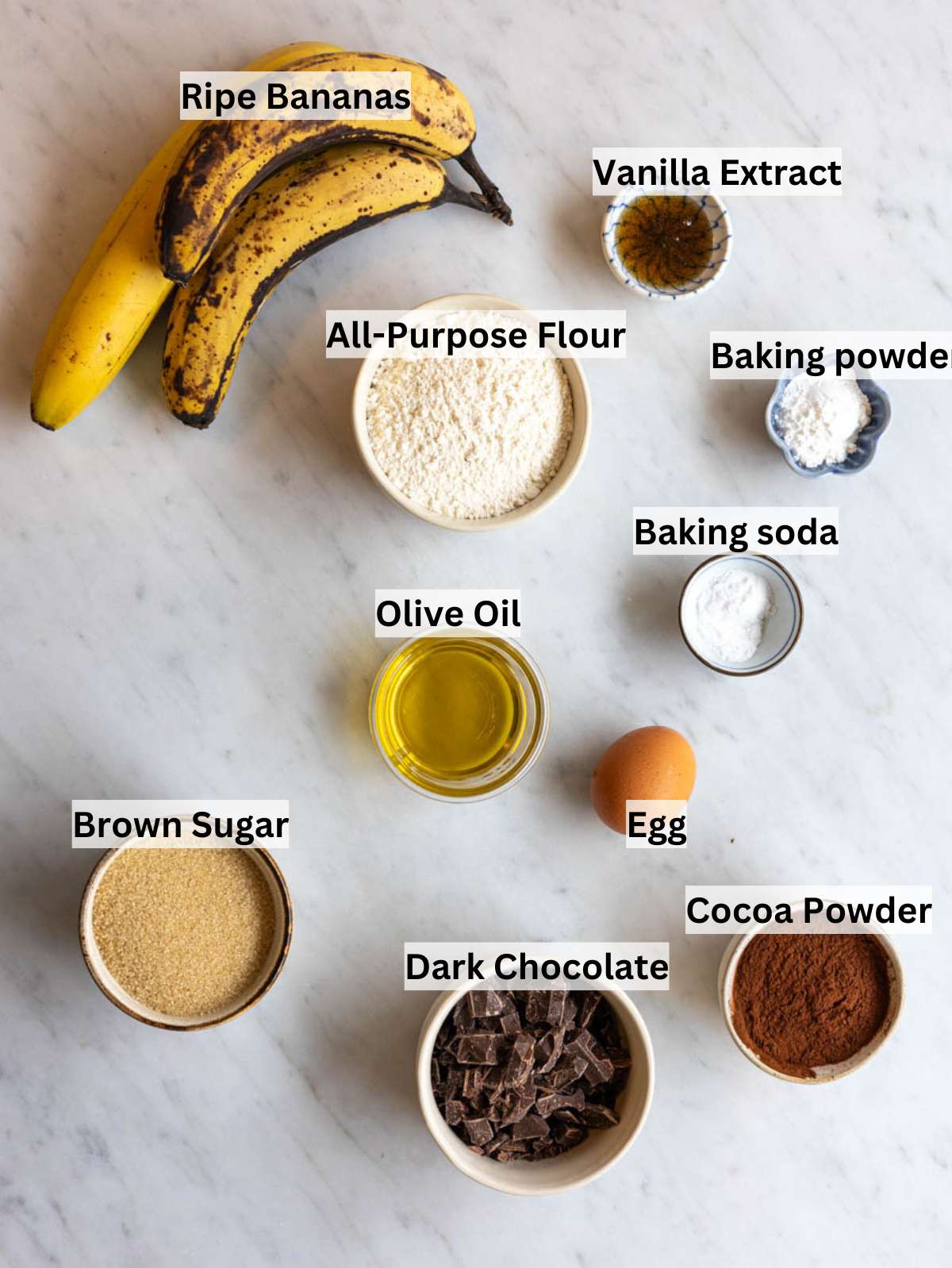 Ripe bananas cocoa powder chopped dark chocolate olive oil sugar egg and flour on a clean counter.