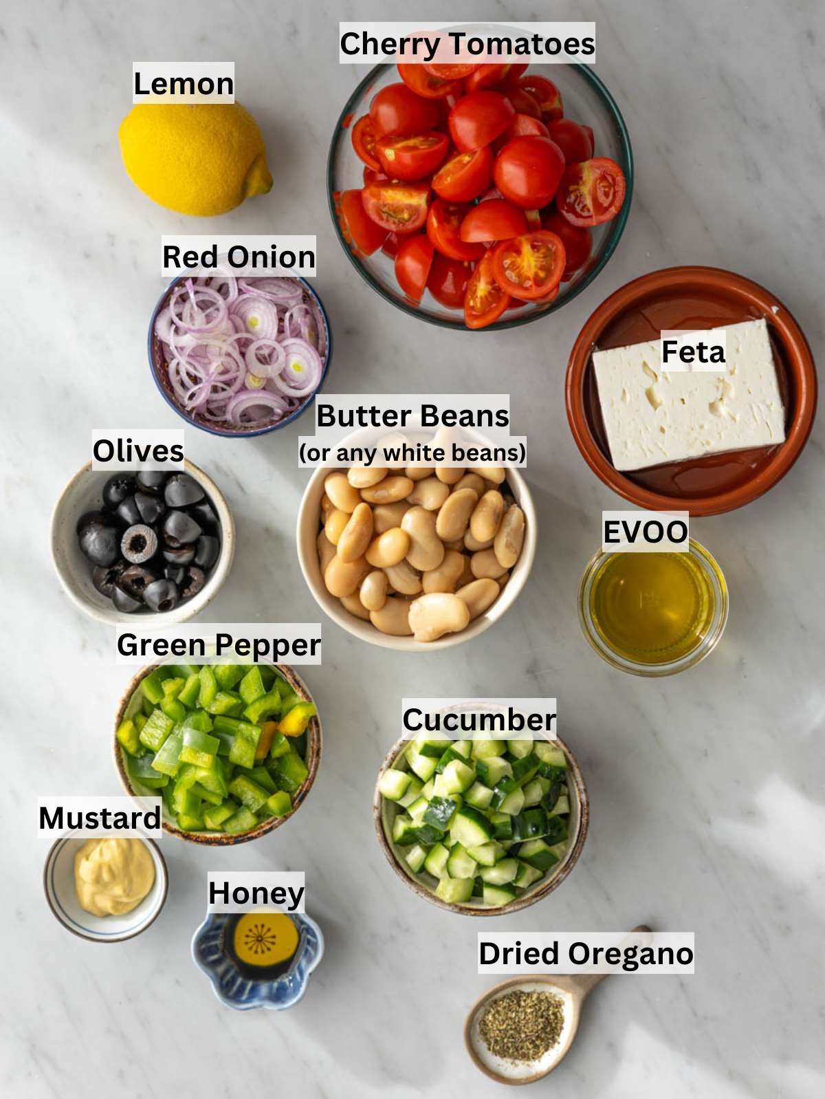 White beans, cucumber, tomatoes, olives, feta, lemon, olive oil arranged on a light kitchen surface.