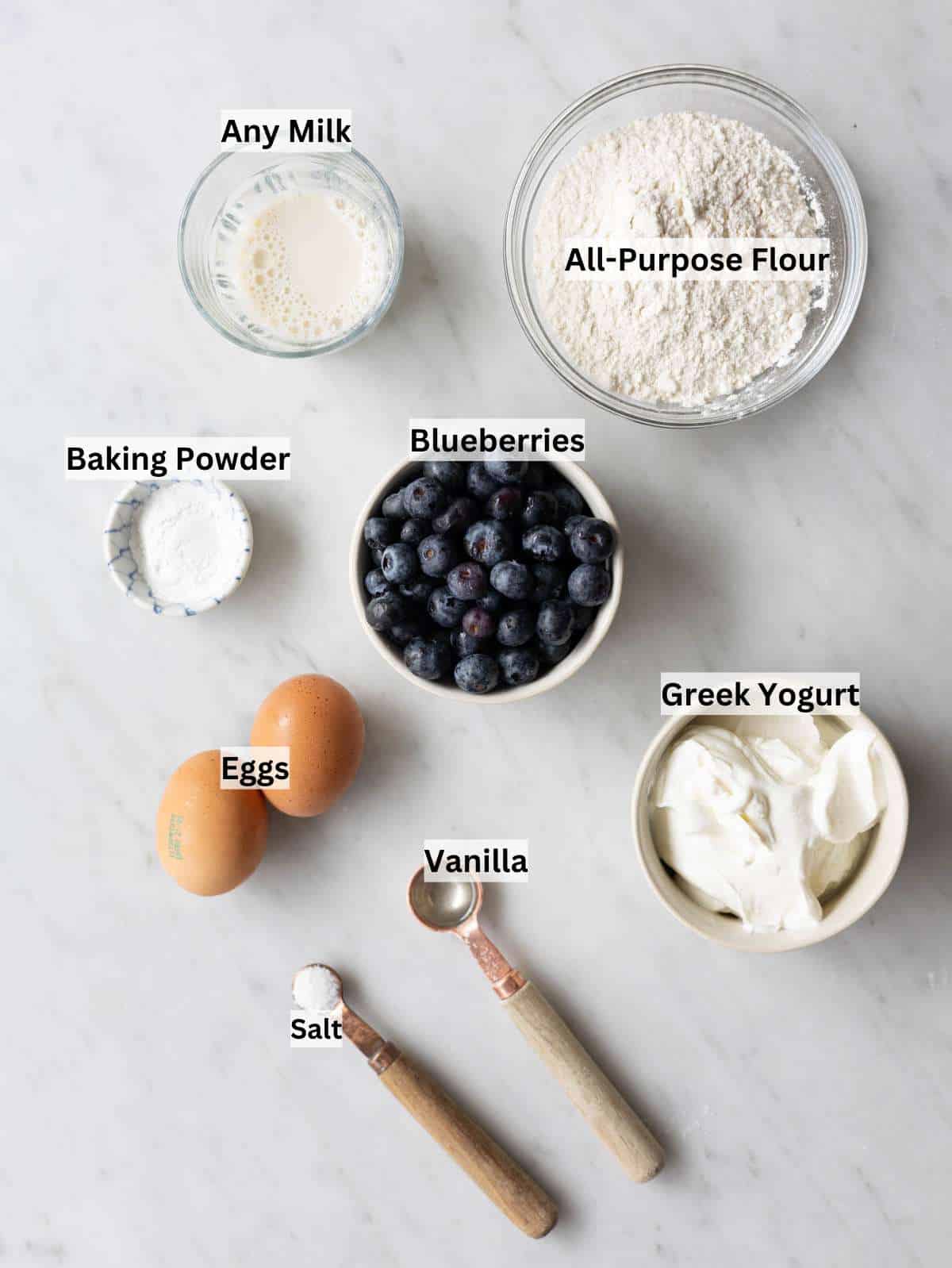 Bowls of flour, Greek yogurt, eggs, milk, and blueberries on marble table.