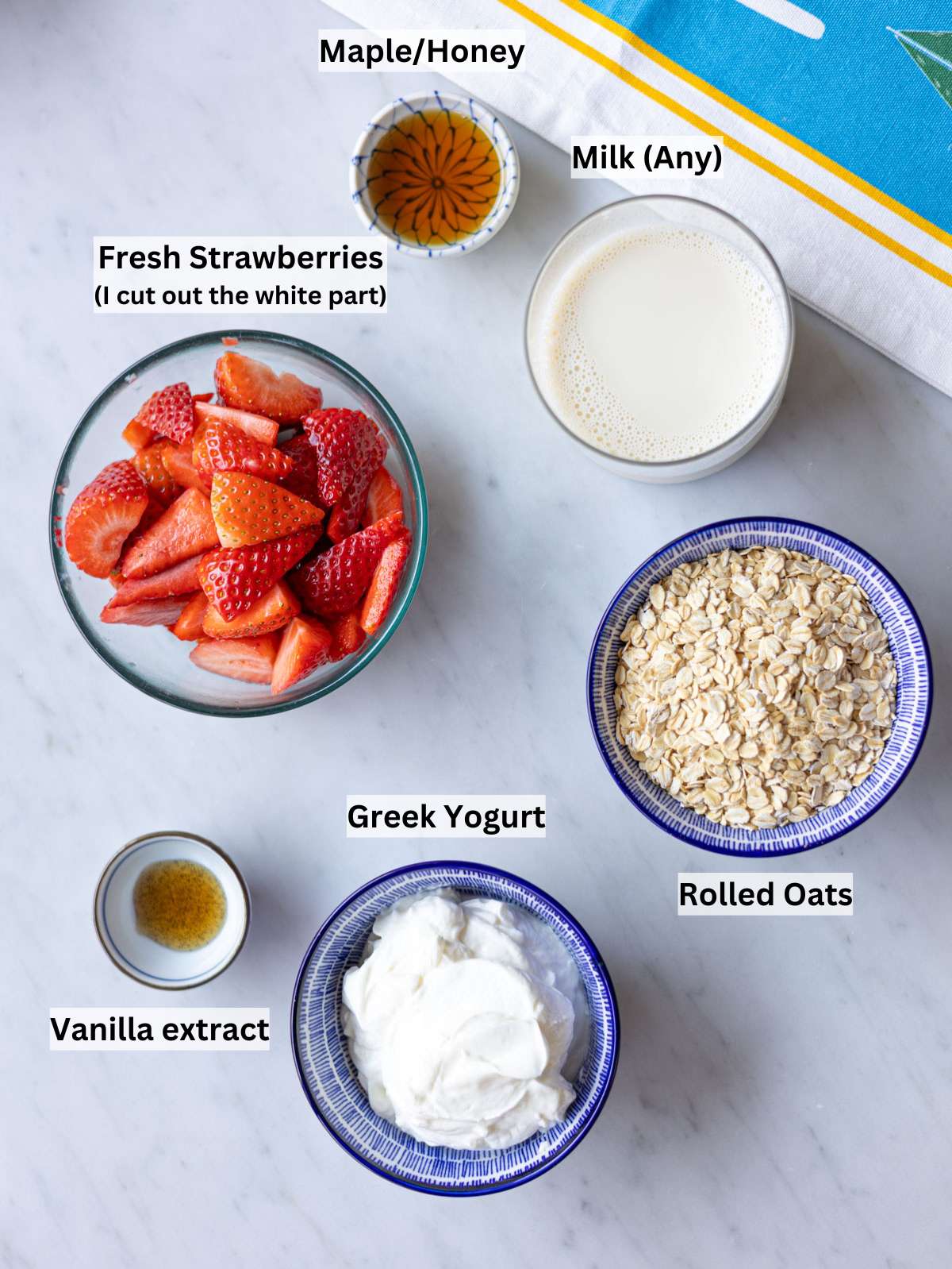 Fresh strawberries, Greek yogurt, milk, maple syrup, vanilla, and rolled oats on table.