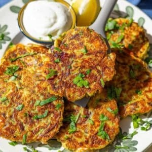 Golden zucchini fritters stacked on plate with creamy dip and herbs.