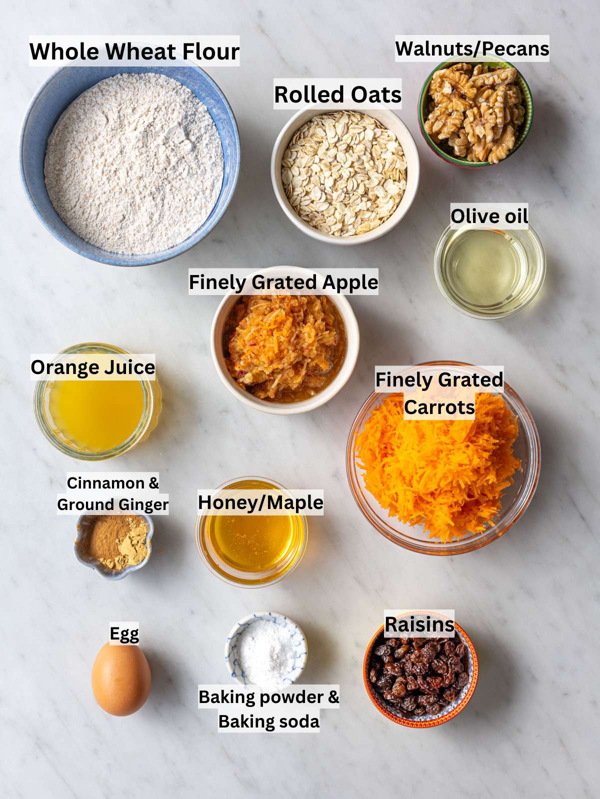 Whole wheat flour, oats, carrots, apple, honey, raisins, walnuts on table.