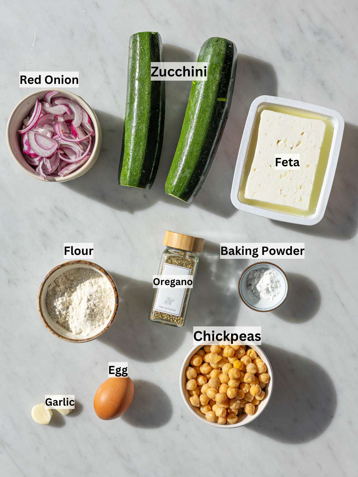 Zucchini, chickpeas, red onion, garlic, feta, flour, and herbs on wooden table.