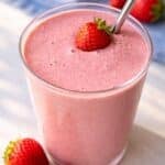 A creamy pink strawberry smoothie in a glass near a sunlit window with marble background.