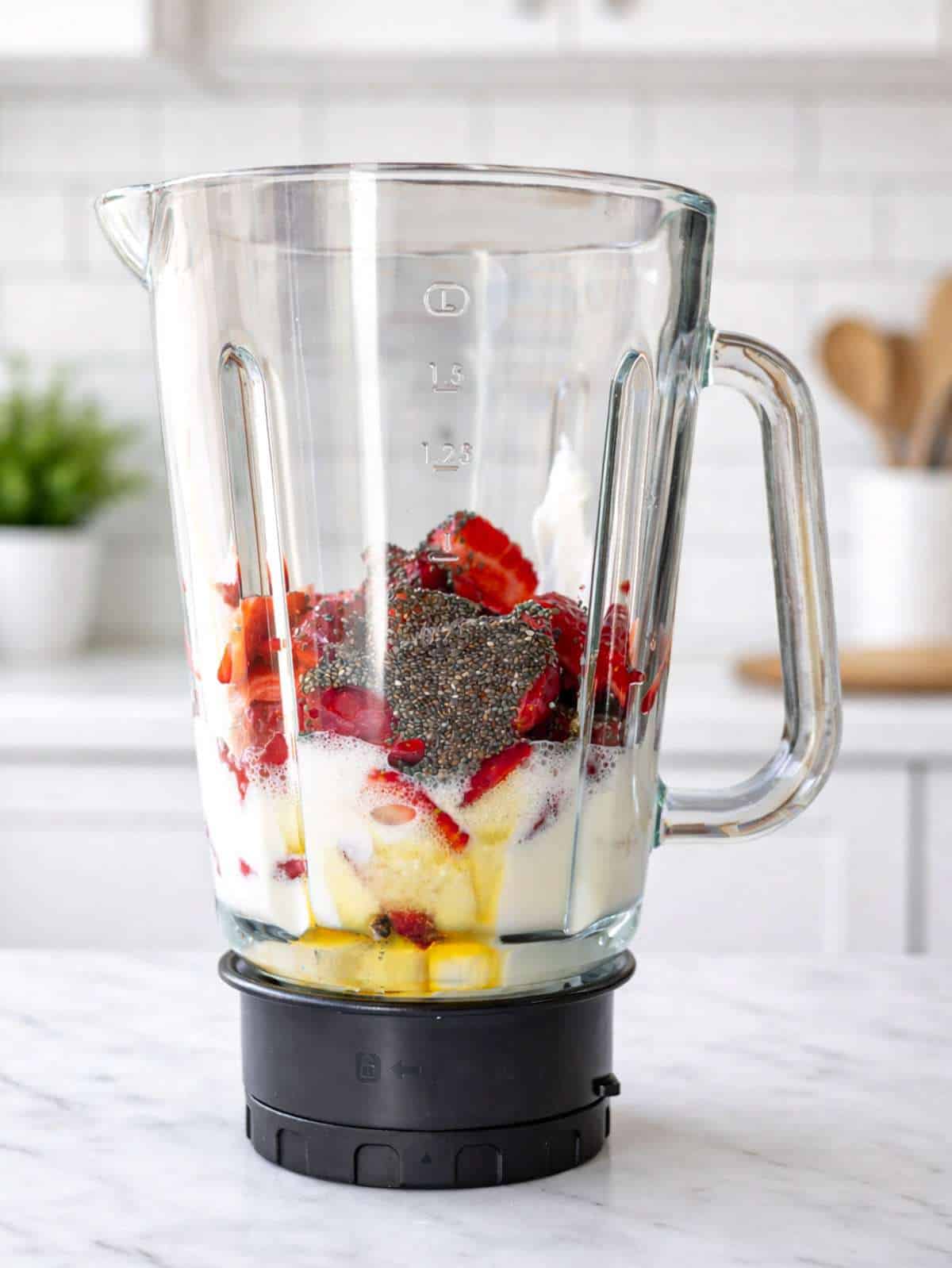A glass blender jar filled with milk, Greek yogurt, strawberries, and chia seeds on marble.