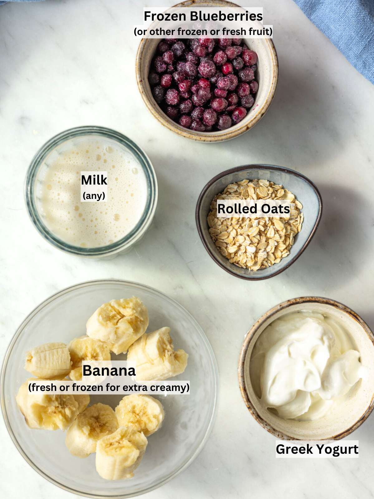 Banana, Greek yogurt, milk, frozen blueberries, oats, and honey arranged on a marble table in natural light.