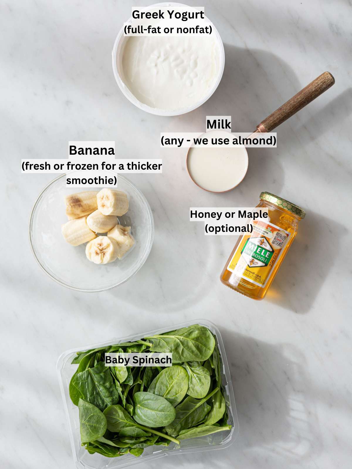 Banana, Greek yogurt, milk, honey, and fresh spinach arranged on marble table in window light.