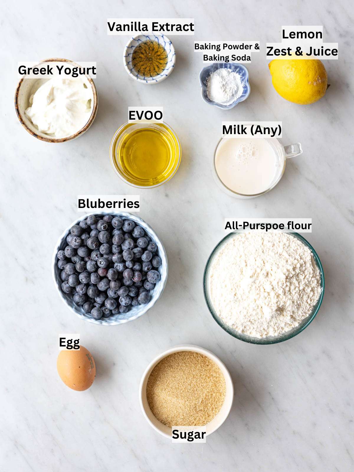 Eggs, Greek yogurt, lemon, blueberries, flour, and sugar on marble surface in soft light.